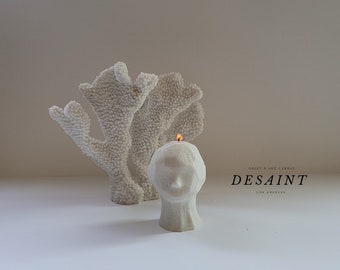 May include: A lit, white, head-shaped candle with a textured surface, next to a white coral sculpture. The candle is labeled "DESAINT LOS ANGELES". The candle's flame is orange. The coral sculpture has a textured surface.