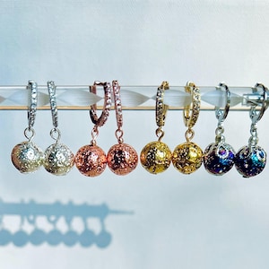 May include: Six pairs of hoop earrings with dangling textured spheres. The hoops are silver, rose gold, gold, and iridescent.