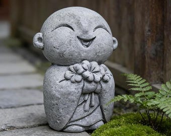 May include: A gray stone statue of a small, smiling figure with a round head and large ears. The figure has closed eyes and an open mouth, holding a cluster of carved flowers against its chest with both hands, featuring a textured surface.