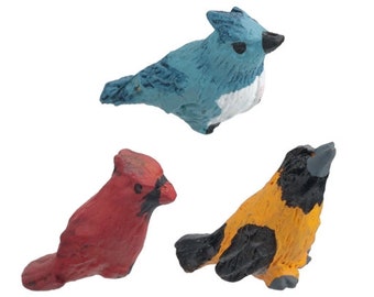 May include: Three small bird figurines, painted in realistic colors. The blue bird has a white belly, the red bird has a black beak, and the yellow bird has black wings.