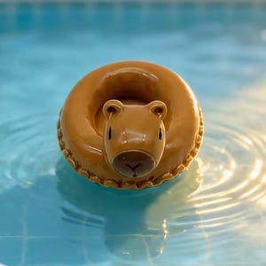 May include: A brown capybara-shaped inflatable pool float. The float is in a ring shape with the capybara's head in the center. The float is floating in a pool of blue water.
