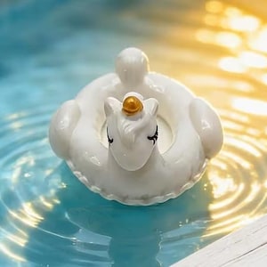 May include: A white ceramic unicorn pool float with a gold horn and black eyelashes floats in a pool. The inflatable ring is round with a small head and tail. The water is blue and reflects sunlight.