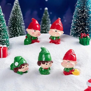May include: Five miniature Christmas elves made of plastic. The elves are wearing red and green hats and outfits. They are standing on a white surface that looks like snow.