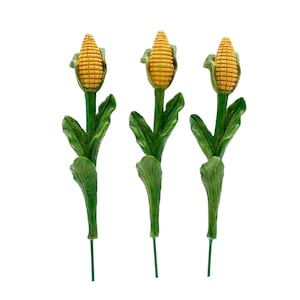 May include: Three artificial corn on the cob stalks with green leaves and yellow corn. The corn is on a white background.