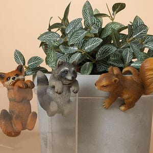 May include: Three small animal figurines, a fox, a raccoon, and a squirrel, are clinging to the sides of three different potted plants. The plants are in gray ceramic pots. The fox is orange and white, the raccoon is gray and black, and the squirrel is brown.