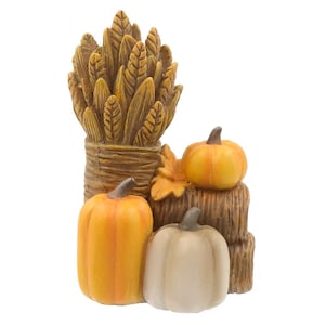 May include: A decorative figurine featuring a tall bundle of brown wheat stalks standing next to a stack of hay bales. Two orange pumpkins and one white pumpkin are positioned at the base, accompanied by a single orange autumn leaf.