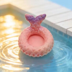May include: A pink and purple mermaid tail-shaped ring float. The float is in a pool of water. The tail is purple at the top and fades to pink. The ring is pink with a textured surface.
