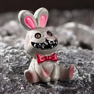 May include: A small, gray rabbit figurine with pink ears and a red bow tie. The rabbit has a wide, toothy grin and large, yellow eyes.