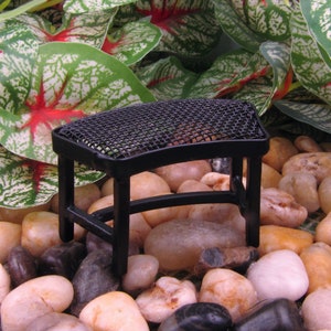 May include: A miniature black metal barbecue grill with a mesh top, set amidst smooth, colorful stones. The grill has a rectangular shape and four legs. Green and white leafy plants are in the background, suggesting a garden or outdoor setting.