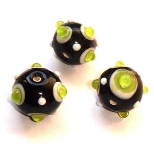 May include: Three black glass beads with white and lime green polka dots. The beads have a textured surface and are designed with a hole in the center for stringing.