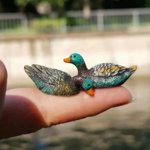 May include: A miniature pair of ducks, featuring teal heads and wings with detailed feathering in shades of brown, green, and yellow. The ducks have orange beaks and are resting on a finger, showcasing their small size and intricate design.