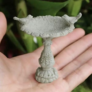 May include: A miniature gray bird bath with two small bird figurines perched on the rim. The bird bath has a detailed pedestal base and a textured basin. The overall design is reminiscent of a classic garden ornament.