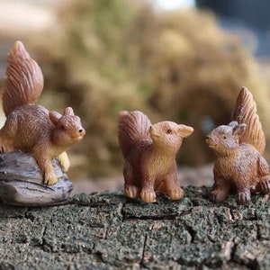May include: Three miniature brown squirrel figurines, two standing on a small rock and one sitting on a log. The squirrels have realistic details and are perfect for a miniature garden or fairy garden.