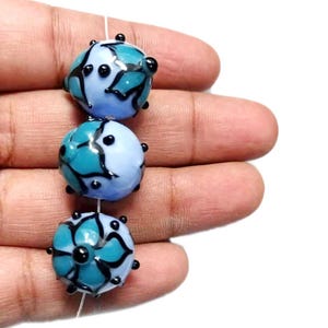May include: Three glass beads in shades of blue and teal, strung on a white thread. Each bead features a floral design in black and teal, with small black accents. The beads are round and appear to be handmade, perfect for jewelry making.
