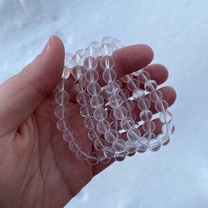 May include: Several clear quartz crystal bead bracelets. Each bracelet is made of round, translucent beads. The bracelets are held in a hand against a blurred, light-colored background. The beads appear to be approximately 8mm in diameter.