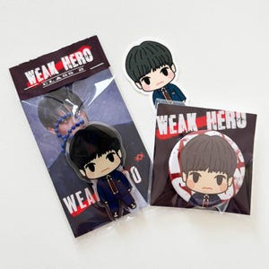 Op de afbeelding: Een Weak Hero Class 2 merchandise set bestaande uit een acryl display in een plastic hoesje, een ronde pin-back button met een bijpassende karakterillustratie en een sticker, allemaal met een donkerharig personage in een pak.
