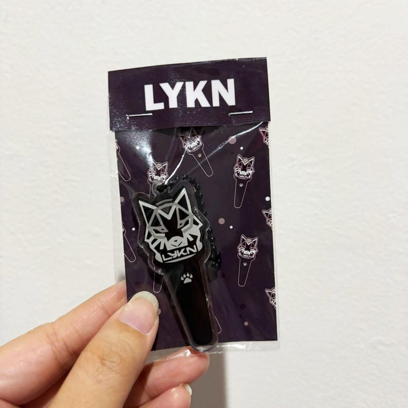 May include: A black and white acrylic keychain shaped like a stylized wolf head with the text LYKN, attached to a black ball chain. The keychain is packaged in a clear plastic sleeve with a dark purple header card featuring the text LYKN.