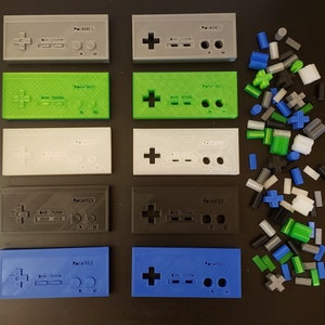 May include: Six different colored 3D printed Nintendo Entertainment System controllers with the text "ReckNES" on each. The controllers are arranged in two rows of three. The controllers are gray, green, white, black, blue, and light blue. There are also a variety of small, colorful plastic pieces scattered around the controllers.