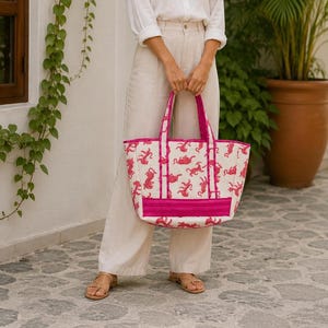Pode incluir: Uma sacola branca e rosa com um padrão repetido de figuras de animais rosa. A sacola tem formato retangular com borda e alças rosa. A sacola é segurada por uma pessoa que usa calças bege e uma camisa branca.