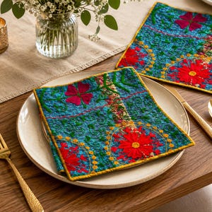Puede incluir: Dos manteles individuales de tela cuadrados con una base azul vibrante y un intrincado bordado floral multicolor en tonos rojo, rosa y amarillo. Cada mantel individual está rematado con un borde amarillo fino y una textura acolchada.