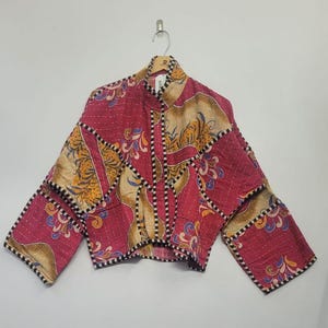 May include: A patchwork jacket in shades of red, gold, and blue, with a high collar and long sleeves. The jacket features a floral pattern and is made of quilted fabric. The jacket is hanging on a hanger.