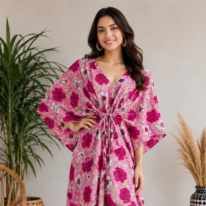 Op de afbeelding: Een roze kaftan-jurk met een levendig bloemenpatroon met donker magenta en witte bloemen. Het kledingstuk heeft een V-hals, wijde kimono-mouwen en een tailleband met trekkoord die de stof in het midden samenbrengt voor een gedrapeerde pasvorm.