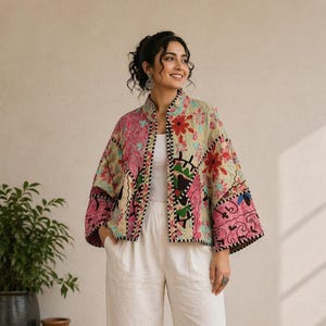 Puede incluir: Chaqueta colorida con estampado floral y cuello alto. La chaqueta presenta un diseño de patchwork con motivos florales rosas, rojos y verdes sobre un fondo beige. Ribete negro perfila los bordes. La modelo lleva pantalones blancos.