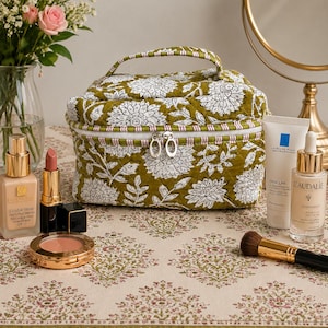 Könnte beinhalten: Eine gesteppte, olivgrüne Kosmetiktasche mit weißem Blumenmuster, einem oberen Griff und einem silbernen Reißverschluss. Make-up-Produkte, darunter Foundation, Lippenstift und Rouge, sind auf einer Oberfläche mit Blumenmuster angeordnet. Ein Blumenstrauß und ein Spiegel befinden sich im Hintergrund.