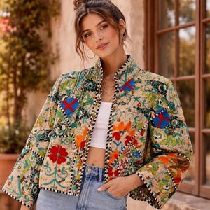 Op de afbeelding: Een kleurrijke gewatteerde jas met bloemen- en geometrische patronen. De jas heeft een beige achtergrond met rode, blauwe, oranje en groene accenten. Het heeft een hoge kraag en een korte pasvorm, gecombineerd met lichtblauwe jeans.