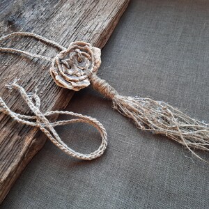 Puede incluir: Un collar hecho a mano con un gran colgante de flor beige texturizado. El colgante está unido a un cordón largo de color marrón con una franja de cuentas beige más pequeñas.