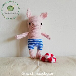 Könnte beinhalten: Eine handgefertigte Häkelpuppe in Form eines Schweins mit rosa Körper und blau-weiß gestreiften Shorts. Das Schwein hat schwarze Augen und kleine Ohren. Neben dem Schwein liegen zwei rot-weiß gestreifte Häkelringe.