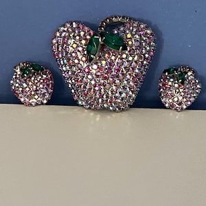 Op de afbeelding: Een vintage sieradenset met een hartvormige broche en bijpassende oorbellen. De stukken zijn ingelegd met iriserende strass in roze, paarse en zilveren tinten. Elk stuk heeft een groene edelsteen accent, wat zorgt voor een glamoureuze uitstraling.