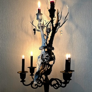 May include: A black metal candelabra with a deer skull in the centre. The candelabra has five candle holders and is decorated with branches and beads. The candles are lit and the room is dimly lit.