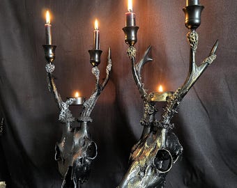 May include: Two black and gold deer skull candle holders with lit candles. The candle holders have a gothic theme. The candles are black and the flames are orange. The candle holders are made of metal.