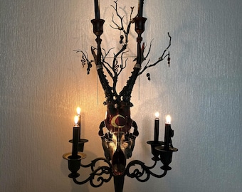 May include: A black metal candelabra with five candle holders. The candelabra is decorated with a deer skull and a tree branch with small ornaments. The candles are lit and the flames are visible.