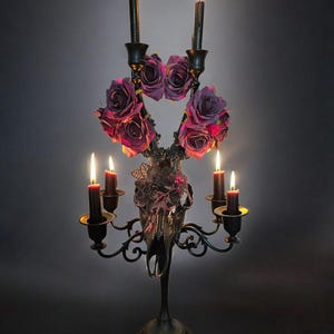 May include: A black metal candelabra with a deer skull base, adorned with purple and pink roses and lit candles.