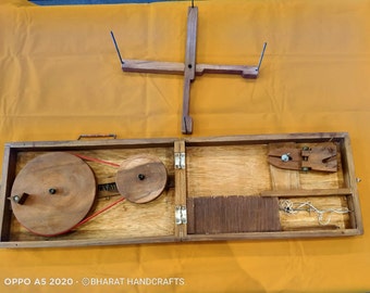 May include: A wooden spinning wheel with a red belt and a wooden frame. The spinning wheel is open and the frame is closed. The spinning wheel is on a yellow surface. The text "OPPO A5 2020 - ©BHARAT HANDCRAFTS" is visible at the bottom of the image.