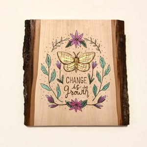 May include: Wooden plaque with a natural edge, featuring a gold butterfly illustration surrounded by leaves and flowers. The text "CHANGE IS GROWTH" is written below the butterfly in black script. The artwork is on a light wood background.