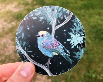 May include: A round sticker featuring a colorful bird perched on a tree branch. The bird has blue, pink, and yellow feathers, set against a dark blue background with stars and stylized foliage. The sticker is held by a hand.