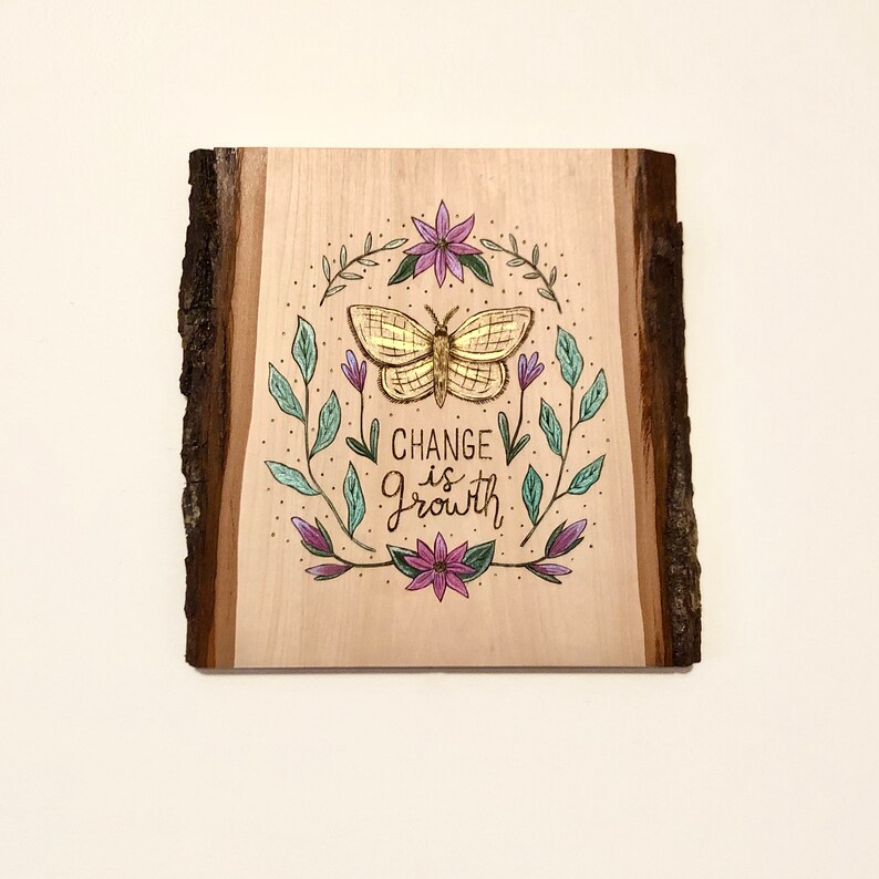 May include: Wooden plaque with a natural edge, featuring a gold butterfly illustration surrounded by leaves and flowers. The text "CHANGE IS GROWTH" is written below the butterfly in black script. The artwork is on a light wood background.