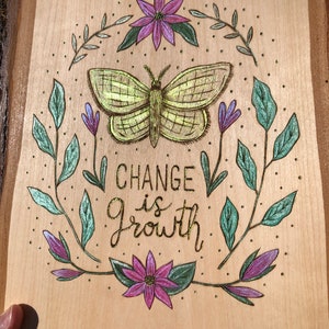 May include: Wooden plaque with a hand-drawn design featuring a central butterfly, surrounded by flowers and leaves. The text "CHANGE is Growth" is written in gold. The artwork is set against a light wood background.
