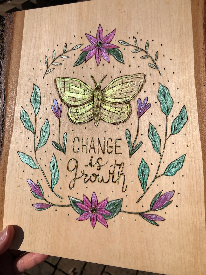 May include: Wooden plaque with a hand-drawn design featuring a central butterfly, surrounded by flowers and leaves. The text "CHANGE is Growth" is written in gold. The artwork is set against a light wood background.