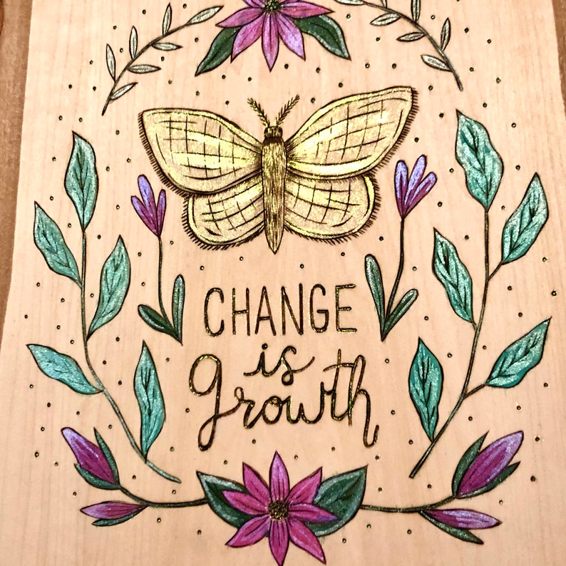 May include: Wooden plaque with a hand-drawn design. A golden butterfly is centered above the words "Change is Growth". The design includes green leaves, purple flowers, and small dots, all on a light wood background.