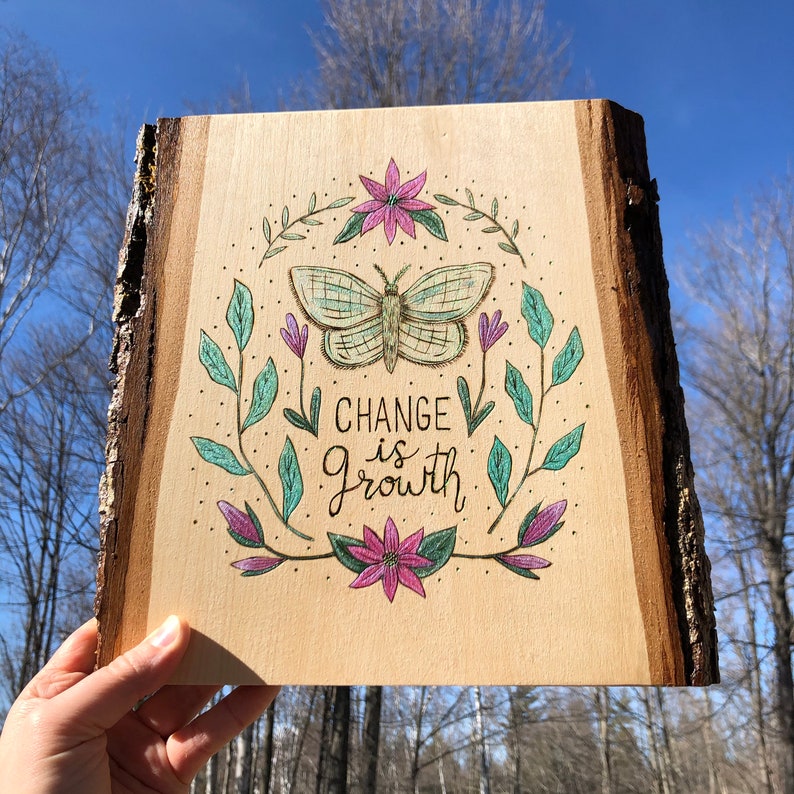 May include: Wooden plaque with a natural edge, featuring a hand-drawn design. The artwork includes a butterfly, floral elements, and the words "CHANGE is Growth" in script. The color palette includes green, purple, and brown.
