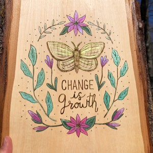 May include: Wooden plaque with a natural edge, featuring a hand-drawn butterfly and floral design. The text "CHANGE is GROWTH" is centered below the butterfly. The artwork is in shades of gold, purple, and teal.