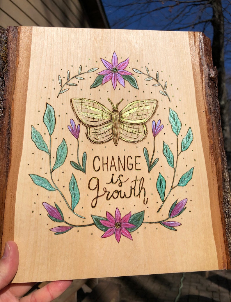 May include: Wooden plaque with a natural edge, featuring a hand-drawn butterfly and floral design. The text "CHANGE is GROWTH" is centered below the butterfly. The artwork is in shades of gold, purple, and teal.