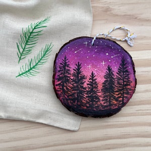 May include: Hand-painted wooden ornament featuring a forest scene under a starry purple and pink sky. The ornament is round with a natural bark edge and a twine hanger. A canvas bag with green painted pine needles is in the background.