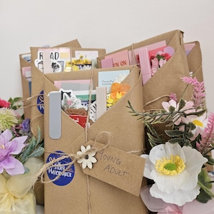May include: Several brown paper wrapped packages tied with twine, each containing books and stationery items. A cardboard tag on the front package is handwritten with the words Young Adult. Decorative artificial flowers are placed nearby.