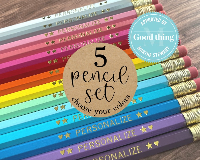 May include: A set of five rainbow colored pencils with gold accents and the words "PERSONALIZE" and "5 pencil set choose your colors" printed on them. The pencils are arranged in a rainbow pattern, with the red pencil at the top and the purple pencil at the bottom. The pencils are on a wooden surface. The pencils are approved by Good Thing Martha Stewart.