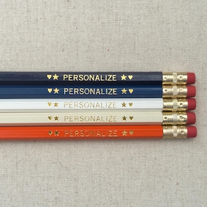 May include: Five personalized pencils with gold stars and hearts. The pencils are navy blue, white, and orange.